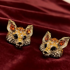May include: A pair of fox-shaped stud earrings crafted from gold-toned metal, featuring intricate detailing encrusted with clear, yellow, and orange crystals. Each fox head has black crystal eyes and teardrop-shaped orange stones in the ears.