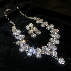 May include: A glamorous silver necklace and earring set. The jewellery features a floral design with numerous clear, sparkling crystals. The necklace and earrings are displayed on a black background, highlighting the brilliance of the crystals.