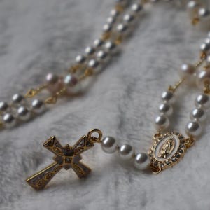 May include: A rosary featuring white spherical beads strung on gold-toned wire, an oval-shaped centerpiece depicting a figure in relief, and a gold-toned cross with intricate detailing and white enamel accents, creating a religious accessory.