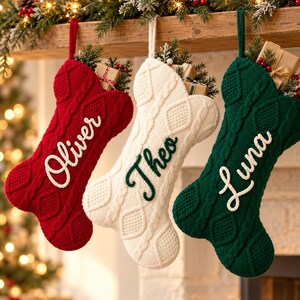 May include: Three bone-shaped Christmas stockings in red, white, and green knit fabric. Each stocking features a cable knit pattern and is embroidered with a name: Oliver in white, Theo in dark green, and Luna in white script lettering.