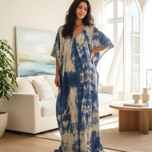May include: A long, flowing dress with a V-neck and short sleeves. The dress features a tie-dye pattern in shades of blue and white. The model is standing in a well-lit room with a couch and a round wooden table.