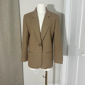 May include: A tan blazer with a single button closure and two front pockets. The blazer is displayed on a mannequin. The blazer has a classic lapel design and long sleeves. The blazer is a neutral color.