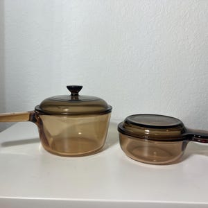 May include: Two amber-colored glass cookware pieces with matching lids. One is a saucepan with a long handle, and the other is a smaller dish with a shorter handle. Both have a classic, vintage design, perfect for cooking and serving.