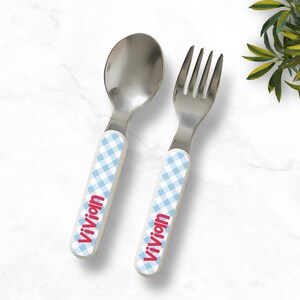 Könnte beinhalten: Ein Löffel- und Gabelset aus Edelstahl mit Griffen in hellblau-weißem Vichy-Karo-Muster. Der Name Vivian ist in roter Schrift auf dem Griff jedes Bestecks aufgedruckt. Das Set ist mit abgerundeten Kanten für Kinder konzipiert.