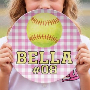 Puede incluir: Un plato redondo con un patrón de cuadros rosa y blanco. El plato presenta una pelota de softbol amarilla con costuras rojas. El nombre "BELLA" y el número "#08" están impresos en negrita, en amarillo.