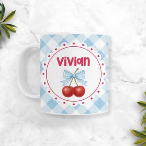 May include: A white ceramic mug featuring a light blue and white gingham pattern. The centre displays a circular scalloped frame with small red hearts, the name Vivian in red text, and an illustration of two red cherries with a blue gingham bow.