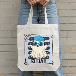 Puede incluir: Una bolsa de tela blanca con el gráfico de un fantasma que lleva una gorra de béisbol azul y gafas de sol redondas. El fantasma está centrado dentro de un marco de rayas azul claro. El nombre ELIJAH aparece impreso en letras mayúsculas.