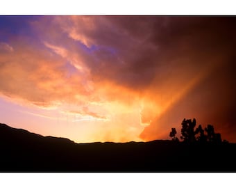 May include: A dramatic sunset with vibrant orange and yellow hues dominates the sky. Dark, silhouetted mountains and trees are in the foreground. The clouds are illuminated by the setting sun, creating a striking contrast.
