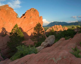 May include: A scenic view of red rock formations in a mountainous landscape. The rocks are illuminated by the setting sun, casting long shadows across the scene.