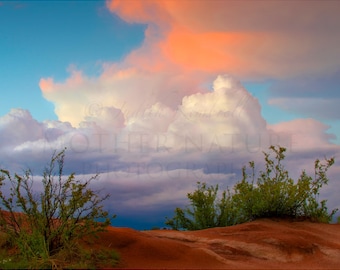 May include: A vibrant sunset sky with fluffy white clouds and a hint of orange, set against a backdrop of red-brown earth and green bushes.