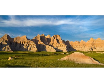 May include: A panoramic view of a grassy plain with a range of tan-colored rock formations in the background. The sky is blue with white clouds.