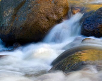 May include: A close-up of a rushing stream of water flowing over smooth, rounded rocks. The water is blurred, creating a sense of motion. The rocks are a mix of brown, gray, and green.