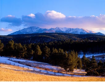 May include: A snow-capped mountain range in the distance, with a forest of evergreen trees in the foreground. The sky is a light blue with white clouds. There is a wooden fence in the foreground, and the ground is covered in snow and dry grass.