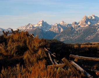 May include: A wooden fence post with a view of snow-capped mountains in the distance. The mountains are in the background and the fence post is in the foreground. The fence post is made of rough-hewn wood and is weathered and worn. The mountains are covered in snow and are a light blue color. The sky is a light blue color and there are clouds in the sky.