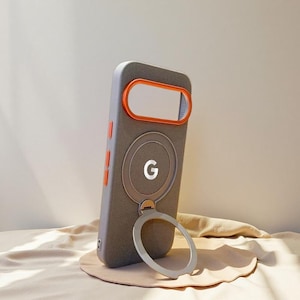 Pode incluir: Uma capa de telefone cinza com detalhes em laranja, com um acessório magnético circular com a letra "G" e um suporte de anel prateado. A capa está sobre uma superfície bege.