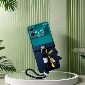 May include: A teal and navy blue phone case with a black wrist strap and a gold-coloured charm. The phone screen displays the time 15:18. The case has a decorative charm in the shape of a bear.