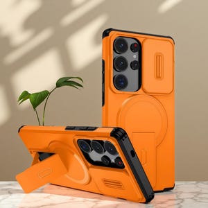 May include: Orange phone case with a black border and a built-in kickstand. The case has a sliding camera cover and a circular magnetic attachment area. The phone case is shown in two positions, one standing upright and the other lying down.