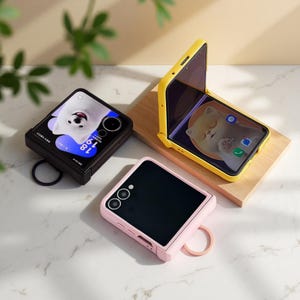 Könnte beinhalten: Drei Smartphone-Hüllen in Schwarz, Gelb und Rosa. Die schwarze Hülle zeigt einen weißen Cartoon-Hund auf dem Bildschirm, während die gelbe Hülle eine Katze zeigt. Die rosa Hülle ist schlicht. Jede Hülle hat einen Ring zum einfachen Tragen.