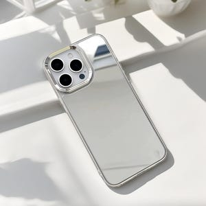 May include: A clear phone case with a reflective, mirrored back. The case has a silver-colored frame and a camera cutout with three black lenses and a flash. The case is designed to fit a smartphone.