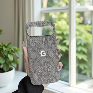 May include: A grey phone case with a snakeskin texture and a silver camera aperture. The case features the Google "G" logo. The case is held in front of a window with foliage.