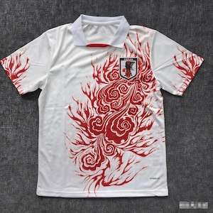 Könnte beinhalten: Weißes Kurzarmtrikot mit rotem, stilisiertem Flammendesign. Das Trikot hat einen weißen Kragen und das Emblem der japanischen Nationalmannschaft auf der Brust. Die Ärmel haben rote Akzente.
