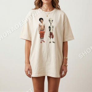May include: Cream-colored t-shirt featuring a graphic of two figures: a person in orange shorts and a towel, and a gray alien in red shorts. The shirt has a relaxed fit.