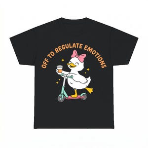 May include: A black t-shirt featuring a graphic of a white duck with a pink bow, riding a teal and pink scooter while holding a coffee cup. The text "OFF TO REGULATE EMOTIONS" is printed in an orange arc above the duck, accompanied by small yellow stars.