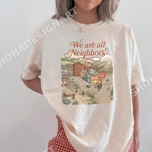 May include: Off-white t-shirt with a town illustration and the text "We are all Neighbors." The graphic shows houses, trees, and cars, with mountains in the background. The shirt has short sleeves and a round neckline.