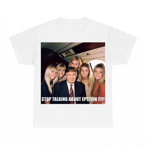 May include: A white t-shirt featuring a graphic print of a man in a dark suit and red tie surrounded by several young women inside an airplane cabin. The text STOP TALKING ABOUT EPSTEIN !!!!! is printed in white capital letters at the bottom.