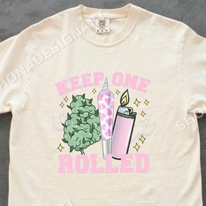 Puede incluir: Camiseta color crema con un diseño gráfico. El diseño incluye el texto "KEEP ONE ROLLED", un cogollo de cannabis verde, un porro con estampado rosa y blanco y un encendedor rosa con una llama, todo delineado con estrellas amarillas.