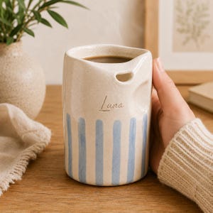May include: A cylindrical ceramic mug with a speckled cream finish, featuring vertical light blue stripes around the base and the name Luna engraved in cursive script. The rim has a distinct molded indentation near the top edge.