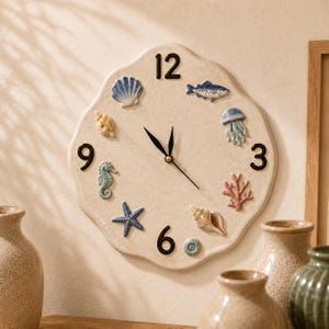 May include: A cream-colored ceramic wall clock with a scalloped edge, featuring black numerals at 3, 6, 9, and 12 o'clock. The clock face is decorated with raised, hand-painted marine life figures including a seashell, fish, jellyfish, seahorse, starfish, and coral.