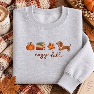 May include: A light gray crewneck sweatshirt featuring an embroidered design of an orange pumpkin, a stack of three books, a maple leaf, and a brown dachshund wearing a scarf, with the text "cozy fall" stitched in brown cursive lettering below.