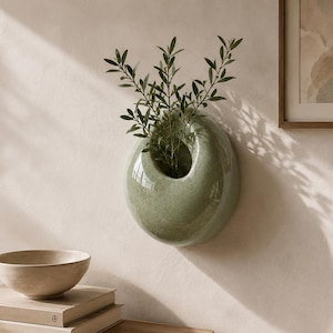 Puede incluir: Una maceta de pared de cerámica en color verde salvia con un acabado liso y moteado y una abertura circular. La maceta contiene una planta verde con tallos delgados y hojas pequeñas y puntiagudas, creando una forma redondeada en la pared.