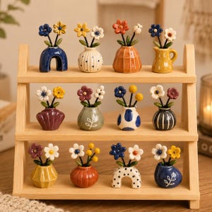 May include: A three-tiered light wood display shelf holding twelve miniature ceramic vases, each containing two small sculpted flowers. The vases feature various shapes, including arches, spheres, and pitchers, decorated with dots, stripes, and solid colors.