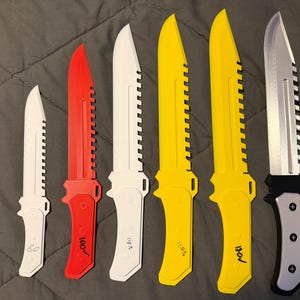 Puede incluir: Seis cuchillos con hojas dentadas en varios colores: blanco, rojo, amarillo y plateado. Algunos cuchillos tienen texto. Los cuchillos están colocados sobre una superficie gris.