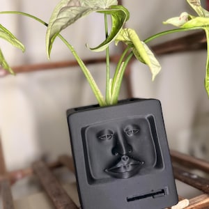 May include: Black square planter with a face design, holding a green plant with large leaves. The planter has a retro computer design. The plant has green and light green leaves. The planter is sitting on a wooden shelf.