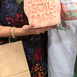 Puede incluir: Una maceta de color melocotón con las palabras "CHUNKY SOIL MIX" en una fuente caprichosa. La maceta tiene forma rectangular con recortes decorativos de hojas y corazones. La imagen es de temática de jardinería.
