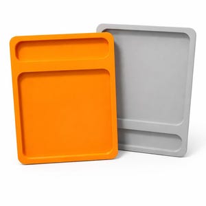 May include: Two rectangular trays, one bright orange and the other grey, are displayed against a white background. Each tray has a recessed area at the top and a slightly raised edge. The trays are designed for storage or organisation.