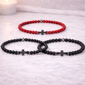 May include: Three beaded bracelets. One red bracelet with a black cross charm and black beads. Two black bracelets with black cross charms and beads. The bracelets are on a white surface.