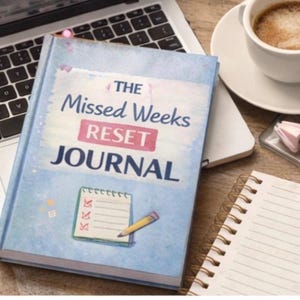May include: A light blue journal titled "The Missed Weeks Reset Journal" with a small illustration of a notepad and pencil. The journal is on a wooden surface next to a laptop, a cup of coffee, and a notepad.