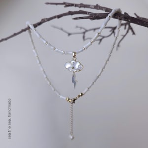 Peut inclure: Un collier à deux rangs avec des perles blanches et des détails dorés. La couche supérieure comporte un pendentif nuage avec un éclair. Le collier est présenté sur une branche. Le texte "sea the sea. handmade" est visible.