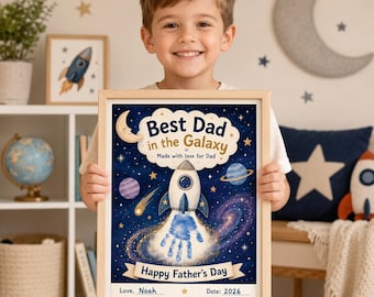 May include: A framed rectangular print featuring a dark blue space theme with stars, planets, and a rocket ship. The text reads Best Dad in the Galaxy, Made with love for Dad, and Happy Father's Day. A blue handprint is centered below the rocket.