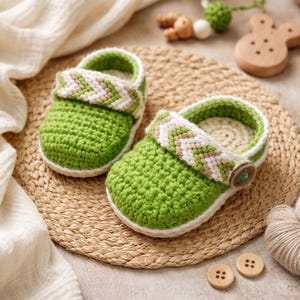 May include: A pair of handmade green crochet baby shoes with a white and tan chevron strap. The shoes have a white sole and a button closure. The shoes are displayed on a woven mat.