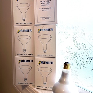 May include: Five white cardboard boxes labeled Premier Reflector Lamp with a line drawing of a light bulb, stacked next to a single BR30 130V 65W reflector lamp with a brass base. The boxes include technical specifications and a manufacture date.