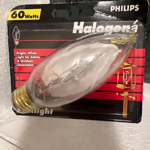 Puede incluir: Un paquete de bombilla Philips Halogena. El paquete es negro y rojo, con el texto "60 Watts" y "Halogena" visibles. La bombilla es transparente con una base dorada. El texto en el paquete dice "Bright, White Light for Safety & Outdoor Decoration."