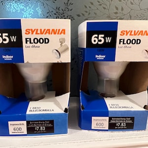 May include: Two Sylvania 65W flood light bulbs in their original packaging. The boxes are white, blue, and brown, with the Sylvania logo and product information printed on them. The boxes also display the bulb's brightness and estimated energy cost.