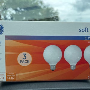 Peut inclure: Un lot de 3 ampoules décoratives G25 blanc doux de la marque GE dans une boîte rectangulaire. L'emballage présente des graphismes orange et bleu, indiquant 40w, 370 lumens et une durée de vie de 1,4 an. La boîte montre trois ampoules rondes.