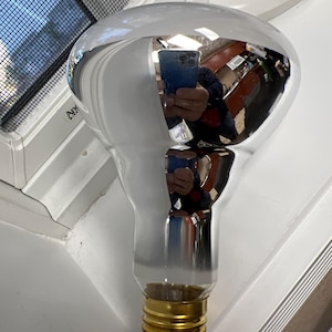 May include: A large, reflective light bulb with a silvered, rounded top and a clear, elongated base. The base has a gold-colored screw-in fitting. The bulb is resting on a white surface near a window.