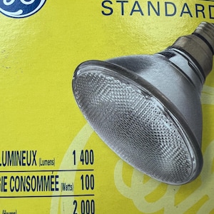 Puede incluir: Una bombilla GE Halogene Standard con un reflector plateado y una lente transparente texturizada. El embalaje es amarillo con texto azul, incluyendo "HALOGENE STANDARD" y especificaciones de salida de luz: 1400 lúmenes, 100 vatios y 2000 horas.